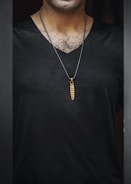 A person wearing a black shirt and a necklace with a pendant resembling a cocoon. The pendant is golden brown and hangs from a beaded chain. The focus is on the torso area, showing some chest hair and part of the neckline.