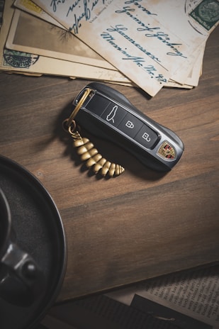 A car key fob with a luxury brand logo is placed on a wooden surface next to vintage letters with cursive handwriting and a golden spiral keychain. The letters have stamps and are partially layered on each other. A section of a dark cup and saucer is visible in the bottom left corner.