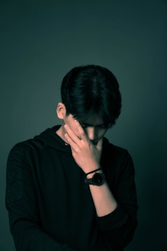 A person wearing a dark hoodie with their hand covering their face, suggesting contemplation or distress. The background is neutral, emphasizing the subdued mood.