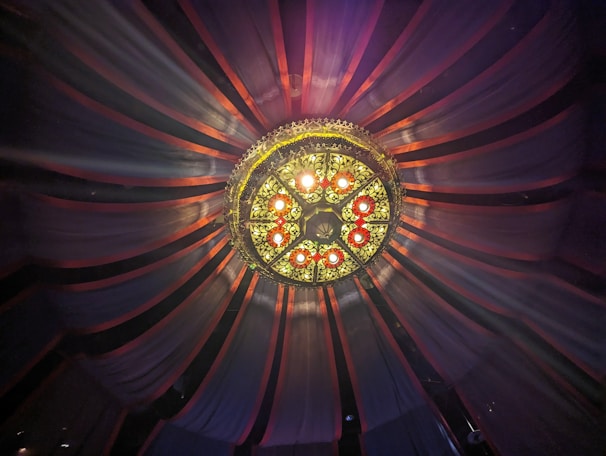 A symmetrical view of an ornate ceiling fixture with a circular pattern of lights and decorative metalwork. The fixture is surrounded by flowing drapery in a radial arrangement extending outward, creating an elegant and theatrical atmosphere.