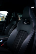 A close-up view of a car interior featuring sleek, dark leather seats with fine stitching and a modern design. A part of the dashboard and center console can be seen, indicating a high-end vehicle.