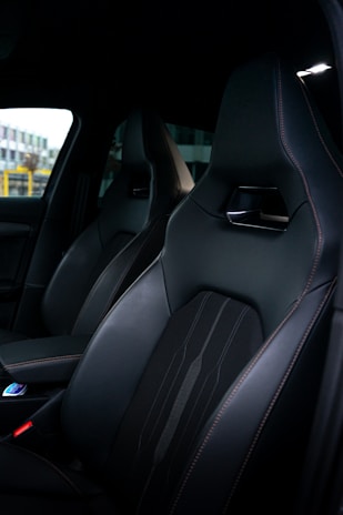 A close-up view of a car interior featuring sleek, dark leather seats with fine stitching and a modern design. A part of the dashboard and center console can be seen, indicating a high-end vehicle.