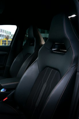 A close-up view of a car interior featuring sleek, dark leather seats with fine stitching and a modern design. A part of the dashboard and center console can be seen, indicating a high-end vehicle.