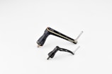 Two metallic bicycle components, including a crank arm and pedal, are placed against a plain white background. The components are sleek and streamlined with a black finish and some silver metallic elements.