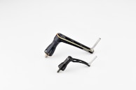 Two metallic bicycle components, including a crank arm and pedal, are placed against a plain white background. The components are sleek and streamlined with a black finish and some silver metallic elements.