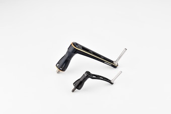 Two metallic bicycle components, including a crank arm and pedal, are placed against a plain white background. The components are sleek and streamlined with a black finish and some silver metallic elements.