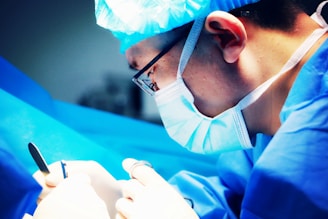 A surgeon wearing a blue surgical mask and cap is intensely focused while performing a procedure. The scene is dominated by shades of blue, and the surgeon is holding surgical instruments.