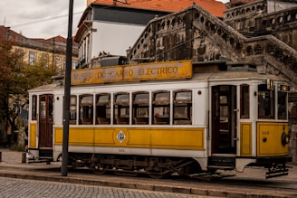 A vintage yellow and white tram is displayed on tracks, next to a cobblestone street. The tram has a sign that reads 'Museu do Carro Eléctrico' and has an old-fashioned design with wooden windows. There is a historic stone building with ornate stairs in the background and other buildings partially visible.
