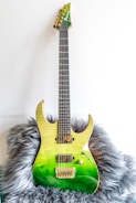 A green electric guitar with a gradient finish from yellow at the top to deep green at the bottom. It has a sleek, modern design and rests on a fluffy gray surface. The guitar features gold hardware, including the bridge and machine heads, and appears well-crafted.