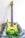 A green electric guitar with a gradient finish from yellow at the top to deep green at the bottom. It has a sleek, modern design and rests on a fluffy gray surface. The guitar features gold hardware, including the bridge and machine heads, and appears well-crafted.