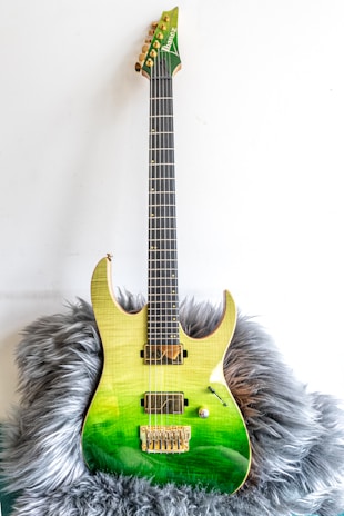 A green electric guitar with a gradient finish from yellow at the top to deep green at the bottom. It has a sleek, modern design and rests on a fluffy gray surface. The guitar features gold hardware, including the bridge and machine heads, and appears well-crafted.