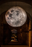 A large, detailed moon sculpture is suspended in an arched corridor. The surface of the moon is illuminated, showcasing craters and lunar textures against the dark surroundings. In the background, there is a glimpse of an outdoor area with trees and lights, adding depth to the scene.