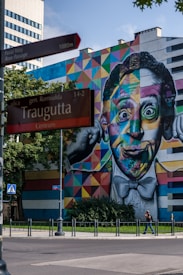 A vibrant mural of a colorful face covers the side of a building with geometric patterns. A street sign indicating Traugutta street is in the foreground, with trees and a lamppost nearby. A person is walking on the sidewalk, and the sky is clear and blue.