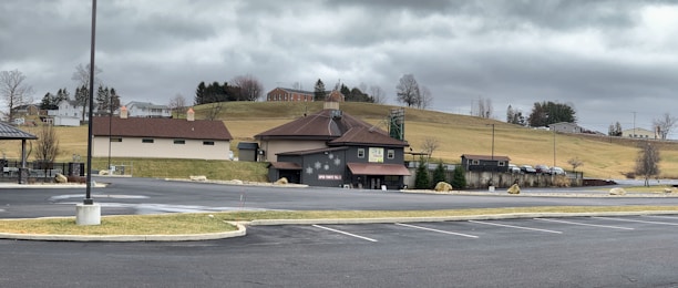 A rural landscape with a large building resembling a barn in the foreground, surrounded by an almost empty parking lot. In the background, grassy hills, scattered trees, and a few residential houses are visible under a cloudy sky.