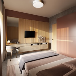A modern bedroom featuring a minimalist design with warm, earthy tones. The room includes a large bed with a neatly arranged blanket and pillows. A sleek wooden dresser is mounted on the wall, supporting a flat-screen TV and decorative items like books and a vase with dried flowers. Natural light pours through a window covered with sheer curtains, casting soft shadows across the room.