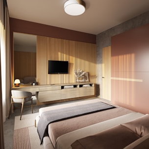 A modern bedroom featuring a minimalist design with warm, earthy tones. The room includes a large bed with a neatly arranged blanket and pillows. A sleek wooden dresser is mounted on the wall, supporting a flat-screen TV and decorative items like books and a vase with dried flowers. Natural light pours through a window covered with sheer curtains, casting soft shadows across the room.