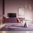 A stylish modern living room features a minimalist design with a round table with a gold-accented base, a plush pink chair, and a large purple carpet. A sleek floor lamp stands to the side, and a console with a flat-screen TV is placed against a soft pink wall. Above the setting hangs a contemporary chandelier, and a framed abstract artwork adds a touch of sophistication.