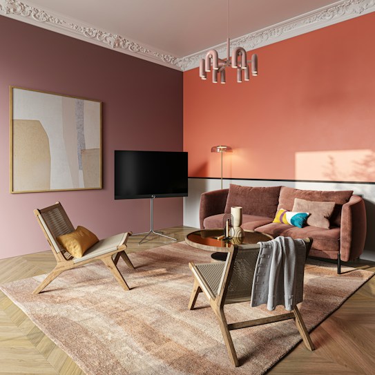 A modern and stylish living room featuring a combination of muted pink and coral walls with intricate ceiling moldings. The room includes a plush velvet sofa with colorful pillows, two wooden chairs with woven seats, and a soft area rug with pastel hues. A sleek black TV is mounted on a stand, and a unique pendant lamp hangs from the ceiling. A round coffee table with decorative items sits in the center, complemented by a floor lamp.