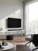 A modern living room with a wall-mounted flat-screen TV above a sleek console. There is a soundbar beneath the TV and a round black speaker on the wooden floor. The room features a large window with sheer white curtains, allowing natural light to enter. A round coffee table with decorative items like a plant and books sits in the foreground next to a comfortable chair with a blanket draped over it.