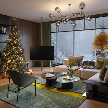A cozy living room with a modern interior, featuring a Christmas tree adorned with lights and a star on top. The room has a large window showing a snowy landscape outside. There is a comfortable, light-colored sofa with decorative cushions, and a uniquely designed chandelier hanging above. The floor is covered with a green rug with yellow accents, and there are a couple of stylish coffee tables with decorative items on them, including a vase with flowers.