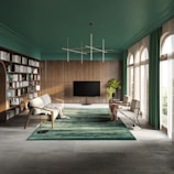 A modern, spacious living room with a sophisticated design. The room features a large bookshelf on the left filled with books and decor items. In the center, a sleek, flat-screen TV stands on a dark stand. Comfortable, stylish chairs and sofas are arranged around a plush green rug. Large windows on the right allow natural light to flood in, highlighting the green indoor plant beside the TV. The ceiling is painted a rich green color, complementing the wooden paneling on the walls and the emerald curtains framing the windows. A contemporary geometric light fixture hangs overhead.
