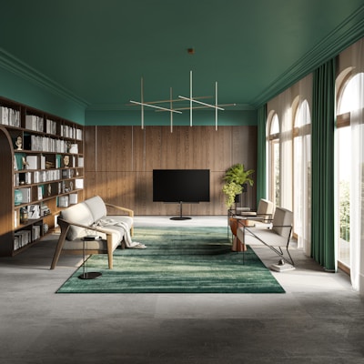 A modern, spacious living room with a sophisticated design. The room features a large bookshelf on the left filled with books and decor items. In the center, a sleek, flat-screen TV stands on a dark stand. Comfortable, stylish chairs and sofas are arranged around a plush green rug. Large windows on the right allow natural light to flood in, highlighting the green indoor plant beside the TV. The ceiling is painted a rich green color, complementing the wooden paneling on the walls and the emerald curtains framing the windows. A contemporary geometric light fixture hangs overhead.