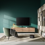 A modern living room setup featuring a large flat-screen television mounted on a dark green wall above a light wood media console. To the left, there is a green upholstered chair with a throw draped over it, positioned on a beige rug. A decorative lamp with an orange shade sits on the console, casting a subtle light. To the right, a shelving unit displays books and decorative items, including a sculpted head.