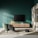 A modern living room setup featuring a large flat-screen television mounted on a dark green wall above a light wood media console. To the left, there is a green upholstered chair with a throw draped over it, positioned on a beige rug. A decorative lamp with an orange shade sits on the console, casting a subtle light. To the right, a shelving unit displays books and decorative items, including a sculpted head.