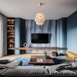 A contemporary living room featuring a large flat-screen TV mounted on a stylish blue and wood-accented wall. A modern chandelier with a geometric design hangs from the ceiling, illuminating the room. The space is furnished with a gray sectional sofa, a wooden coffee table adorned with books, and decorative glassware. Shelving units hold various books and decorative items, while a vibrant blue rug adds color to the wooden floor.