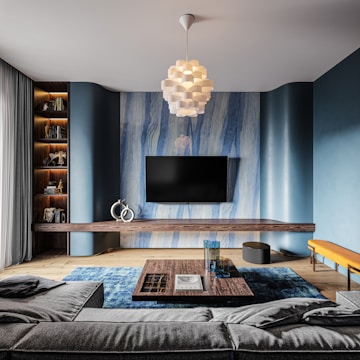 A contemporary living room featuring a large flat-screen TV mounted on a stylish blue and wood-accented wall. A modern chandelier with a geometric design hangs from the ceiling, illuminating the room. The space is furnished with a gray sectional sofa, a wooden coffee table adorned with books, and decorative glassware. Shelving units hold various books and decorative items, while a vibrant blue rug adds color to the wooden floor.