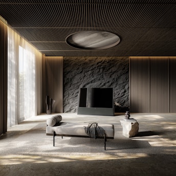 A modern, minimalist interior featuring a room with a textured stone wall and a sleek, black television. A daybed with a throw blanket is positioned centrally on a woven rug. To the right, a small stone table holds decorative objects, and sheer curtains allow soft natural light to filter through.