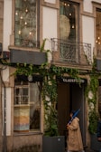 A storefront with large windows partially covered by creeper plants. The facade features decorative metal balconies and exterior lighting. One person wearing a beige trench coat and holding a blue umbrella is walking past the shop, which has the name 'CLAUS PORTO' displayed above the entrance.