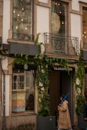 A storefront with large windows partially covered by creeper plants. The facade features decorative metal balconies and exterior lighting. One person wearing a beige trench coat and holding a blue umbrella is walking past the shop, which has the name 'CLAUS PORTO' displayed above the entrance.