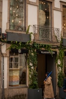 A storefront with large windows partially covered by creeper plants. The facade features decorative metal balconies and exterior lighting. One person wearing a beige trench coat and holding a blue umbrella is walking past the shop, which has the name 'CLAUS PORTO' displayed above the entrance.