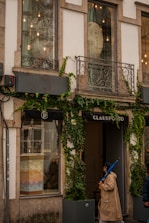 A storefront with large windows partially covered by creeper plants. The facade features decorative metal balconies and exterior lighting. One person wearing a beige trench coat and holding a blue umbrella is walking past the shop, which has the name 'CLAUS PORTO' displayed above the entrance.