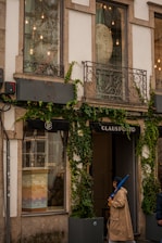 A storefront with large windows partially covered by creeper plants. The facade features decorative metal balconies and exterior lighting. One person wearing a beige trench coat and holding a blue umbrella is walking past the shop, which has the name 'CLAUS PORTO' displayed above the entrance.