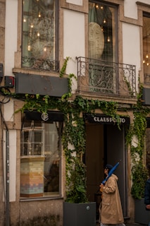 A storefront with large windows partially covered by creeper plants. The facade features decorative metal balconies and exterior lighting. One person wearing a beige trench coat and holding a blue umbrella is walking past the shop, which has the name 'CLAUS PORTO' displayed above the entrance.