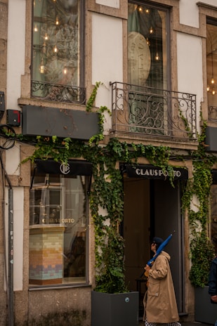 A storefront with large windows partially covered by creeper plants. The facade features decorative metal balconies and exterior lighting. One person wearing a beige trench coat and holding a blue umbrella is walking past the shop, which has the name 'CLAUS PORTO' displayed above the entrance.