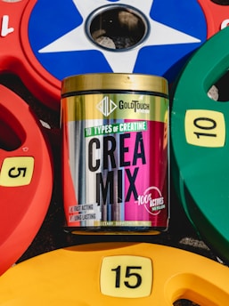 A container of GoldTouch Nutrition's Crea Mix supplement is positioned among colorful weight plates. The container features vibrant colors and text highlighting '10 types of creatine' with claims of being fast-acting and long-lasting. The weight plates are numbered 5, 10, and 15 in yellow, red, green, and blue colors and are arranged around the supplement container.