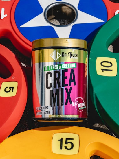 A container of GoldTouch Nutrition's Crea Mix supplement is positioned among colorful weight plates. The container features vibrant colors and text highlighting '10 types of creatine' with claims of being fast-acting and long-lasting. The weight plates are numbered 5, 10, and 15 in yellow, red, green, and blue colors and are arranged around the supplement container.