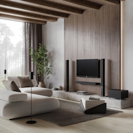 A modern living room with minimalist design, featuring a large, comfortable white sofa and a sleek, wall-mounted television. The room is accented with wooden beams on the ceiling and a large, natural light window that brightens the space. There is a potted plant adding a touch of greenery, and various black audio equipment including speakers are neatly arranged around the TV. A soft gray rug complements the light wooden floor.