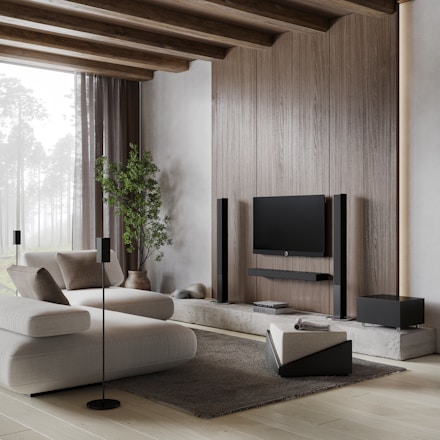 A modern living room with minimalist design, featuring a large, comfortable white sofa and a sleek, wall-mounted television. The room is accented with wooden beams on the ceiling and a large, natural light window that brightens the space. There is a potted plant adding a touch of greenery, and various black audio equipment including speakers are neatly arranged around the TV. A soft gray rug complements the light wooden floor.