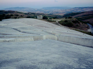 A vast landscape featuring hilly terrain covered in terraces of concrete structures. In the distance, a solitary, worn-down building stands amid the rolling hills. The sky is hazy, with muted colors enhancing the tranquil and somewhat desolate atmosphere.