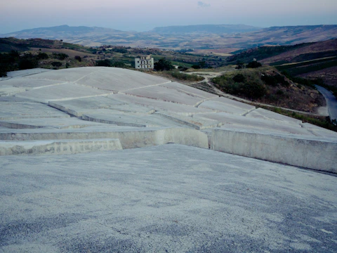 A vast landscape featuring hilly terrain covered in terraces of concrete structures. In the distance, a solitary, worn-down building stands amid the rolling hills. The sky is hazy, with muted colors enhancing the tranquil and somewhat desolate atmosphere.