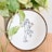 An embroidery hoop displays a minimalist line art of a couple embracing, with added red and green floral accents. The hoop is placed in front of a green potted plant, and rests on a wooden surface.