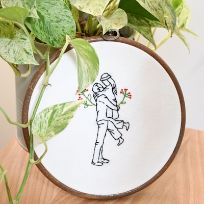 An embroidery hoop displays a minimalist line art of a couple embracing, with added red and green floral accents. The hoop is placed in front of a green potted plant, and rests on a wooden surface.