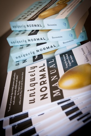 A stack of books titled 'uniquely NORMAL' by Bernstein is neatly arranged in two piles. One pile has books with blue covers, and the other has white and black covers with the word 'MANUAL' prominently displayed.
