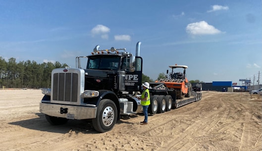 A large black semi-truck with a chrome grille is parked on a sandy construction site. The truck is towing a flatbed trailer carrying an orange heavy-duty construction vehicle. A person wearing a white hard hat and yellow safety vest stands next to the truck. The site is surrounded by dirt and some construction equipment, with trees and blue sky in the background.