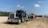 A large black semi-truck with a chrome grille is parked on a sandy construction site. The truck is towing a flatbed trailer carrying an orange heavy-duty construction vehicle. A person wearing a white hard hat and yellow safety vest stands next to the truck. The site is surrounded by dirt and some construction equipment, with trees and blue sky in the background.