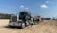 A large black semi-truck with a chrome grille is parked on a sandy construction site. The truck is towing a flatbed trailer carrying an orange heavy-duty construction vehicle. A person wearing a white hard hat and yellow safety vest stands next to the truck. The site is surrounded by dirt and some construction equipment, with trees and blue sky in the background.